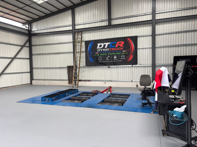 Dinamómetro de chasis azul con dos pares de rodillos, plataforma de acero, estación de control con monitor a la derecha y rótulo de Dyno Truck Costa Rica con los íconos de potencia, torque, consumo y diagnóstico.