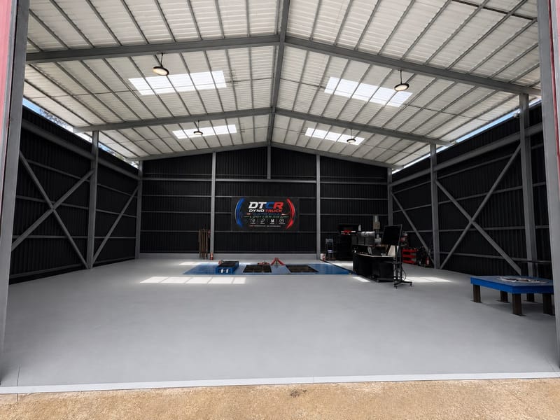 Nave de Dyno Truck Costa Rica vista desde el portón: estructura metálica con techo claro, piso gris, el dinamómetro azul empotrado al fondo, la estación de control a la derecha y el rótulo DTCR Dyno Truck en la pared.