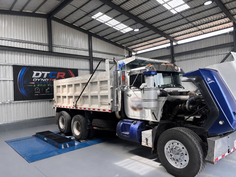 Camión volquete con el capó abierto y los ejes traseros en el dinamómetro, dentro de la nave con el rótulo de Dyno Truck Costa Rica.